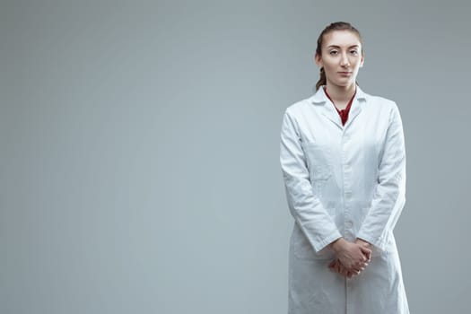 Young confident female technician wearing lab coat is posing in a studio with a serious facial expression, looking directly at the camera with her hands clasped in front of her