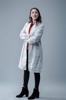 Young confident female technician wearing a lab coat poses with crossed arms in a studio, looking directly at the camera with a serious, determined expression