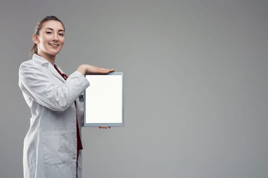Young female doctor wearing lab coat and stethoscope is showing a digital tablet with blank white screen while standing on a gray background