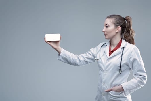 Young female doctor wearing a lab coat and stethoscope, presenting a smartphone with a blank white screen, highlighting a medical app or website for patient engagement and telehealth services