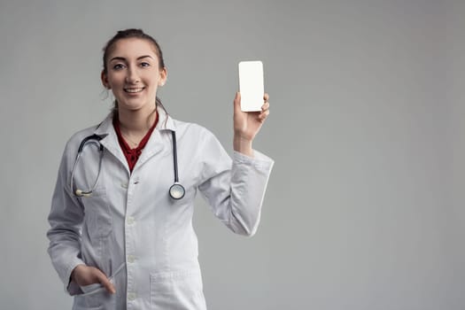 Smiling young female doctor wearing lab coat and stethoscope is showing a smartphone with blank screen, promoting an app or website for healthcare
