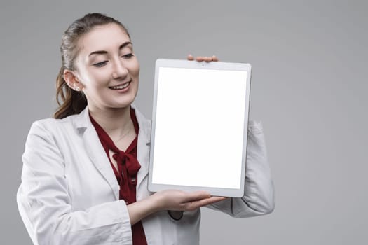 Young female doctor wearing lab coat and stethoscope holding digital tablet with blank screen, looking at it and smiling on plain background