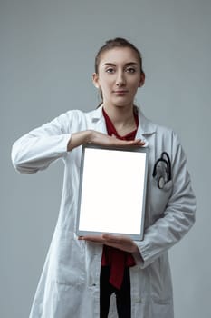 Young female doctor wearing a white coat and stethoscope is holding a digital tablet with a blank screen, providing ample space for custom content related to healthcare and technology