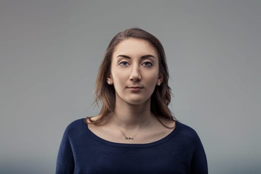 Portrait of a serious young woman with long brown hair and blue sweater staring intensely at the camera against a gray background, conveying confidence and determination
