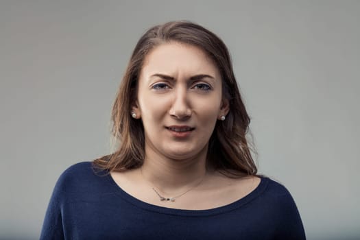 Young woman with long brown hair, wearing a dark blue sweater and necklace, frowning and wrinkling her nose in skepticism against a gray background