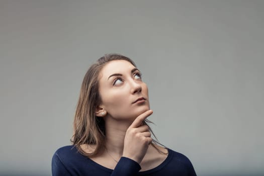 Young woman with long brown hair is thoughtfully looking up with her hand on her chin, contemplating something in a studio setting with a gray background
