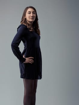Young woman with long brown hair, wearing a dark dress and tights, stands in profile against a gray backdrop, her hand resting confidently on her hip while looking directly at the camera