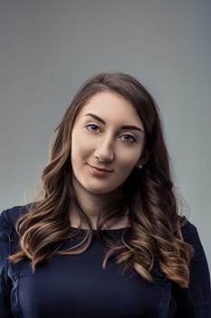 Studio portrait of a young woman with long brown hair smiling confidently against a gray backdrop, conveying a sense of poise and self-assurance