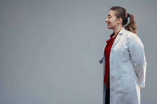 Side view of a young female doctor wearing a white coat and stethoscope, standing with her hands behind her back and smiling warmly, conveying confidence and professionalism in healthcare