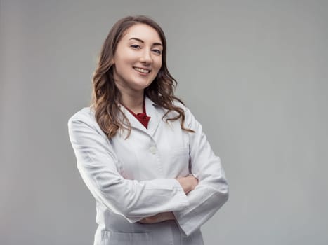 Cheerful young female scientist wearing a lab coat, standing confidently with arms crossed against a gray background, exuding professionalism and expertise