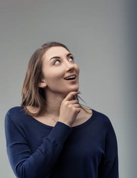 Pensive young woman gazing upwards with a smile, resting her hand on her chin, contemplating future projects and feeling a surge of inspiration, hope, and ambition