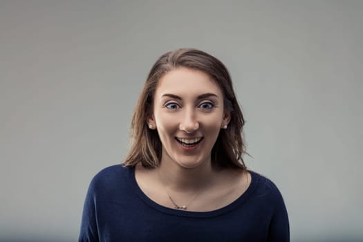 Surprised young woman gasping and smiling with joy, radiating excitement and happiness against a light gray background, showcasing her cheerful expression and vibrant energy