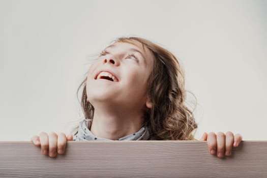 Portrait of a cheerful girl with long brown hair, hiding behind a wooden board and looking up with a happy expression, showing curiosity and joy