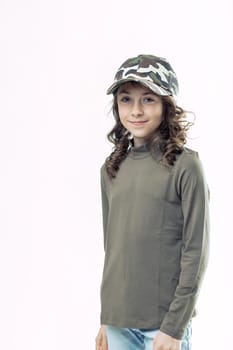 Smiling preteen girl with long brown hair, wearing a camouflage baseball cap, olive green turtleneck, and light blue jeans, posing confidently against a white background