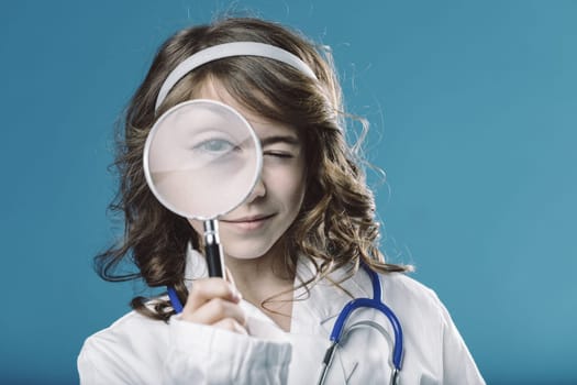 Curious young aspiring doctor winking while looking through a magnifying glass, wearing a white coat and stethoscope against a vibrant blue backdrop