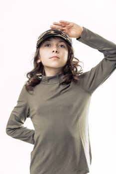 Portrait of a confident young girl standing against a white background, wearing a camouflage cap and saluting with her hand on her forehead, exuding a sense of pride and determination