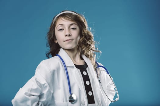 Portrait of a confident young girl wearing a doctors coat and stethoscope, standing against a vibrant blue backdrop with her hands on her hips