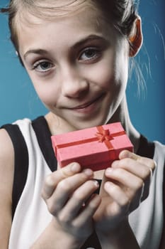 Young girl smiling while holding a small, red gift box wrapped in ribbon against a soft blue background, capturing the joy and spirit of celebration