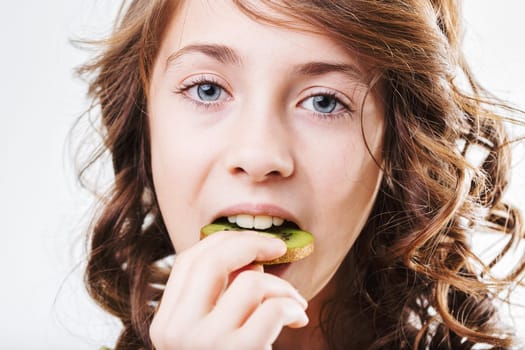 Portrait of a cute little girl with long, curly brown hair enjoying a slice of fresh kiwi fruit while looking directly at the camera against a clean white background