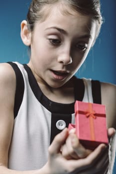 Young girl holds a small, red gift box in her hands, her eyes wide with surprise and curiosity as she opens it against a vibrant blue background
