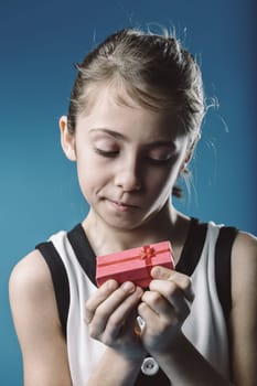 Young girl looking with curiosity at a small pink gift box with a red ribbon, wondering about its content, against a vibrant blue background