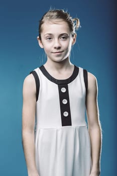 Studio portrait of a smiling young girl wearing a modern white dress with black details, standing against a vibrant blue backdrop, exuding confidence and style