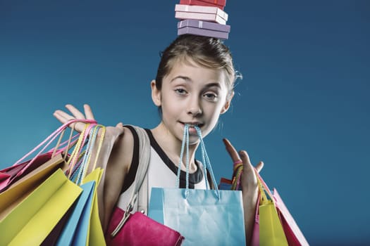 Young girl skillfully balances a stack of gift boxes on her head while juggling numerous shopping bags, embodying the spirit of consumerism against a vibrant blue backdrop