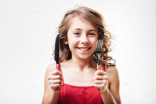 Smiling young girl with wavy hair holds a fork and knife, radiating excitement and joy while eagerly anticipating a delicious meal