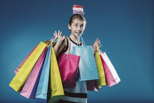 Young girl skillfully balances a stack of colorful gift boxes on her head while juggling numerous shopping bags, radiating happiness and excitement after a successful shopping spree