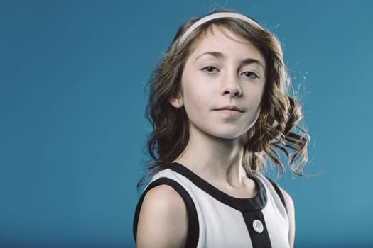 Studio portrait of a smiling young girl with brown curly hair and a white headband, wearing a black and white dress, posing against a vibrant blue backdrop