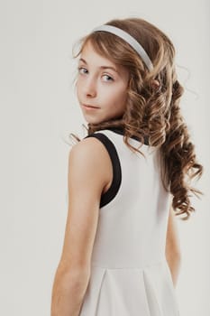 Elementary age girl with long, curly hair and a headband, wearing a sleeveless dress, gazing over her shoulder with a calm expression against a neutral background