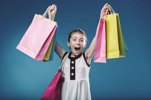 Studio shot of a cheerful elementary age girl holding colorful shopping bags, expressing her excitement after a successful shopping spree, against a vibrant blue backdrop
