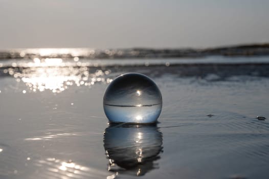 A glass ball lies in the waves on the sandy beach, the sea and the setting sun are reflected in the ball
