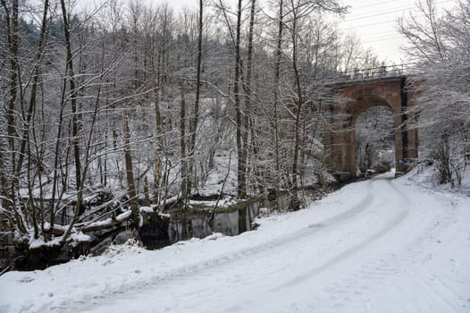 Narrow snow-covered road with a small stream and a viaduct in a landscape with trees