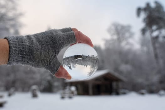 Human hand with a grey glove holds a glass ball reflecting a snow-covered landscape and a hut