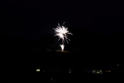 A firework rocket in the sky on a mountain at night, a few illuminated houses in the foreground