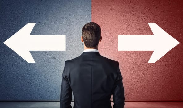 Rear view doubtful businessman in a suit standing in front of a wall divided into two colors, blue on the left and red on the right, with arrows pointing different directions to choose