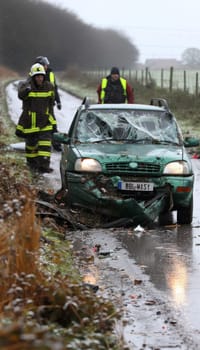 Two emergency responders in high-visibility gear examine a damaged vehicle on a snowy rural road, with the windshield shattered from an apparent collision