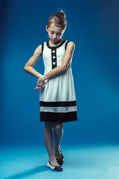 Studio portrait of a shy elementary age girl looking down, wearing a black and white dress and ballet flats, standing on a blue seamless background