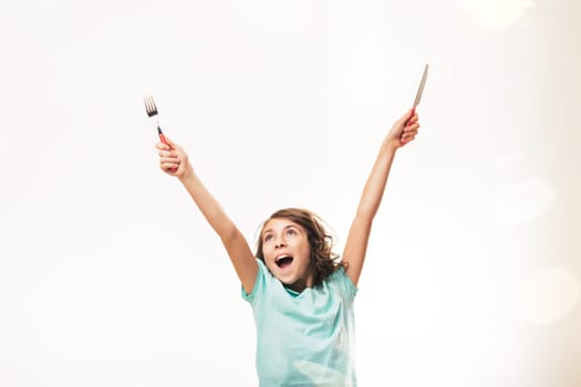 Enthusiastic preteen girl raising her arms, holding a fork and knife, expressing excitement and anticipation for a meal, against a bright white background