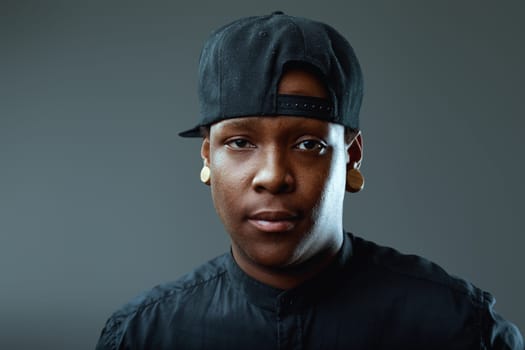 Portrait featuring a serious young african-american man with ear plugs and a baseball cap, staring intensely at the camera against a moody gray background, exuding confidence and style