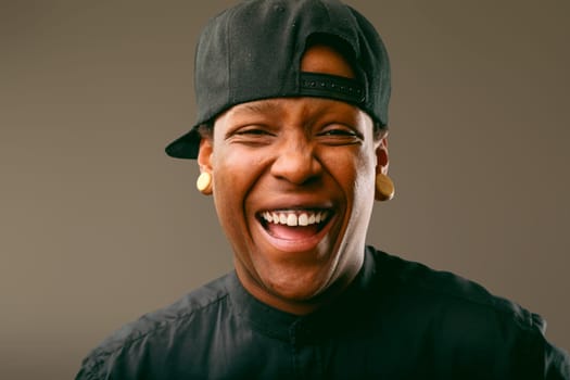 Cheerful young musician wearing a stylish black outfit and baseball cap, laughing joyfully and displaying a bright smile, radiating happiness and positive energy in a studio setting