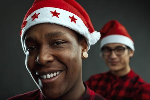 Two diverse young men wearing santa claus hats, smiling brightly and looking directly at the camera during a lively office christmas party, radiating joy and festive cheer