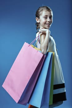 Studio shot of a smiling girl carrying colorful shopping bags on her shoulder against a vibrant blue background, enjoying her successful shopping spree