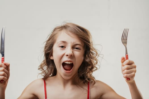 Young caucasian girl with brown curly hair holding a fork and knife, shouting with excitement against a white background, clearly expressing hunger and eagerness to eat