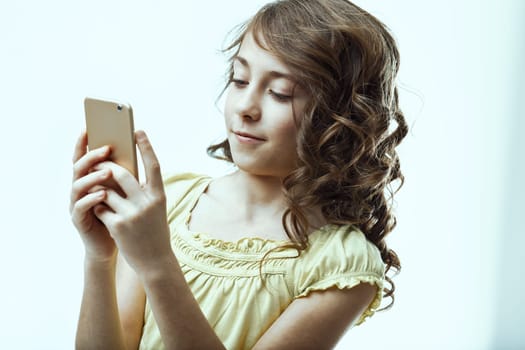 Cute elementary age girl with curly hair, smiling while holding and using a smartphone, engaged with the screen. Bright expression radiates happiness in a studio setting