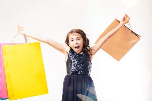 Excited elementary age girl shouting with joy while holding colorful shopping bags, arms outstretched in celebration, isolated against a clean white background