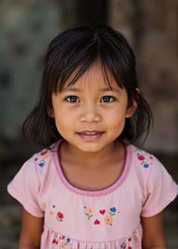 A young girl living in Thailand stands outdoors, her dark hair framing her cheerful face. She wears a pink shirt adorned with colorful flowers, reflecting joy and innocence.
