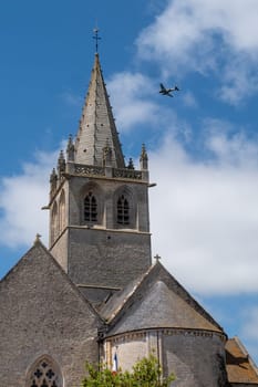 A church steeple with a plane flying in the sky. The plane is flying low and is in the background. Saint Come du Mont, normandy, France