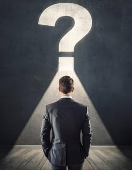 Rear view puzzled businessman in a dark room in front of a wall with illuminated interrogation mark. Finding answer to his question. Contemplation concept